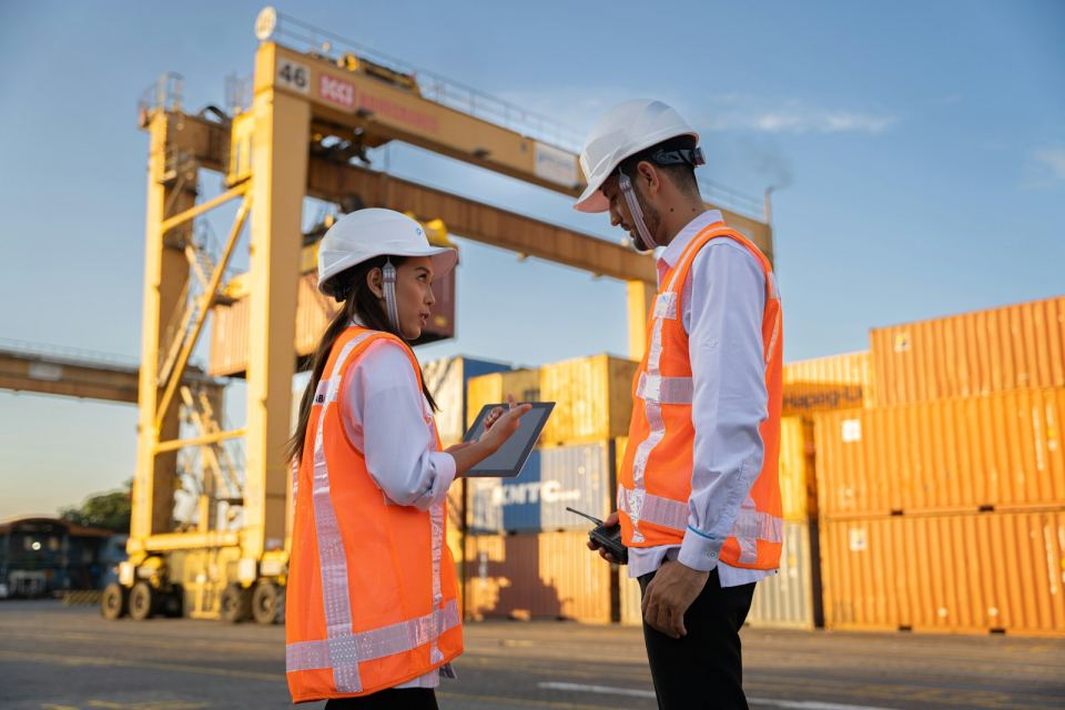 Two workers in hard hats reviewing plans beside a container stack